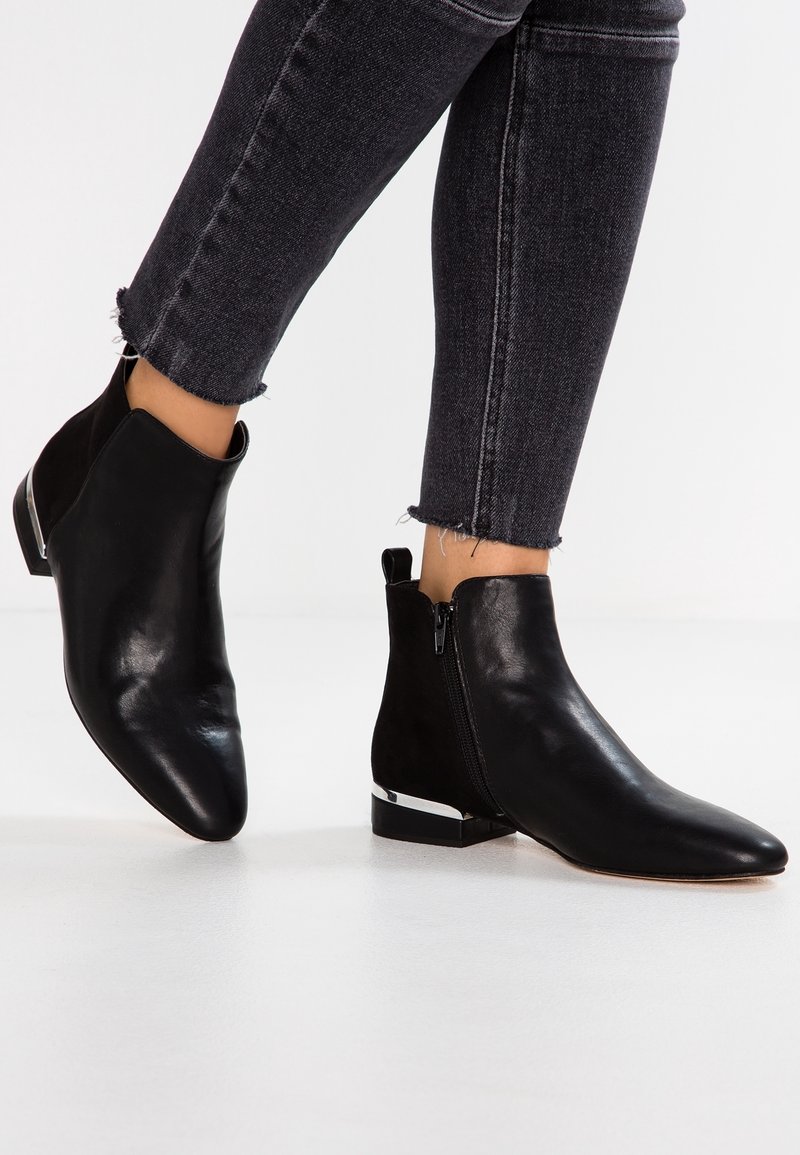 Personne portant des bottines en cuir noir avec des fermetures éclair latérales et un détail argenté au talon, associées à un jean denim noir raccourci.