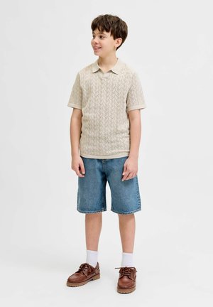 Chłopiec stoi i się uśmiecha, ma na sobie beżową koszulę z krótkim rękawem w wzory, krótkie dżinsowe spodenki do kolan, białe skarpetki i brązowe buty.