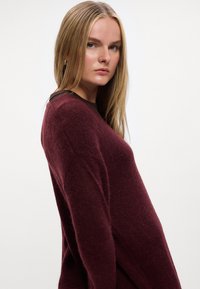 Burgundi gyapjú pulóver hosszú ujjal, laza fazonban, texturált felületű, és kontrasztos barna dekoltázzsal. Oldalnézet, egyszerű háttér előtt.