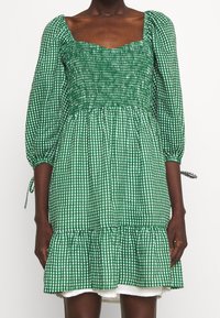 Zelené kostkované šaty se čtvercovým výstřihem, smockovaným tělem, naducanými rukávy a volánkovým lemem. Zobrazeno na modelce s tmavou pletí.