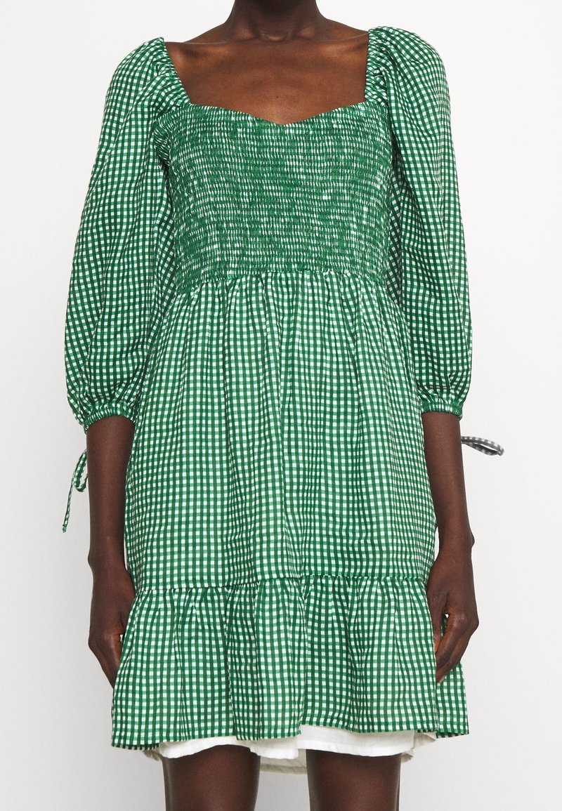 Zelené kostkované šaty se čtvercovým výstřihem, smockovaným tělem, naducanými rukávy a volánkovým lemem. Zobrazeno na modelce s tmavou pletí.