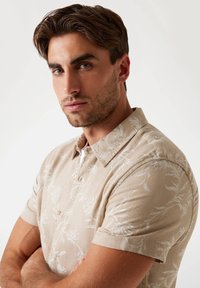 Camisa de manga corta en color beige, confeccionada con una tela ligera, que presenta un patrón botánico blanco y un cuello con botones.