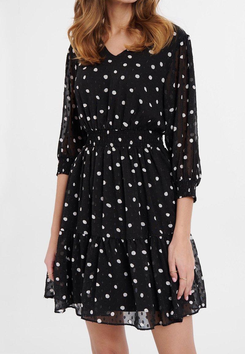 Femme portant une robe noire longueur genou à pois blancs, manches trois-quarts transparentes et taille élastique, debout sur fond blanc.
