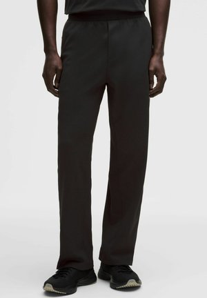 Mężczyzna ubrany w czarne spodnie o prostych nogawkach i czarne trampki stoi na jasnym, jednolitym tle.