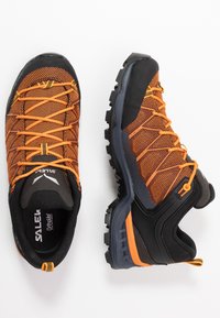 Sporty turskoer med oransje mesh-overdel, svarte gummidetaljer og robust såle. Funksjoner inkluderer snøresystem og trekkstang for enkel påkledning.