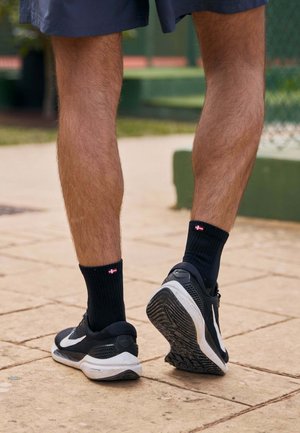 Schwarze Sportschuhe mit weißen Akzenten, getragen mit knielangen schwarzen gerippten Socken, die ein rot-wei�ßes Logo zeigen, von hinten betrachtet.