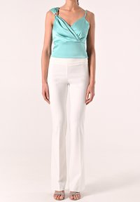 Femme portant un top en satin vert menthe à épaule dénudée avec un détail en anneau doré et un pantalon droit taille haute blanc, associé à des sandales argentées.