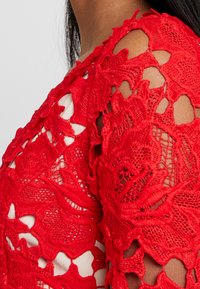 Red crochet lace top featuring an open shoulder design, intricate floral patterns, and a soft inner lining. Textured fabric details are prominent.