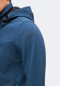 Gros plan d’une personne portant une veste à capuche bleue avec un petit patch rectangulaire sur le haut du bras indiquant ICEPEAK.