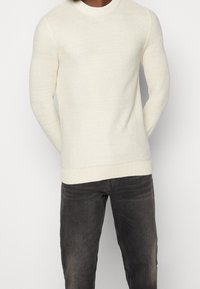 Krémově zbarvený pletený svetr s texturovaným vzorem, kulatým výstřihem a žebrovanými manžetami; v kombinaci s tmavě šedými džínami.