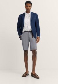 Marineblazer, weißes Hemd, karierten Shorts in Blau und Weiß, brauer Gürtel und braune Slipper. Glatter Stoff, taillierte Passform und kontrastierende Texturen.