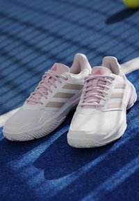 Par hvide og lyserøde sportssko med tre beige striber på en blå tennisbane nær en hvid linje og en tennisbold.