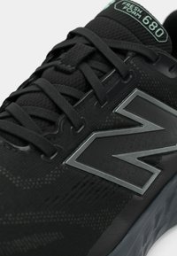Tuva tuvumā melna sporta apava ar teksturētu audumu, melnām auklām, sudraba logotipu sānos un "Fresh Foam 680" uzlīmi uz mēles.