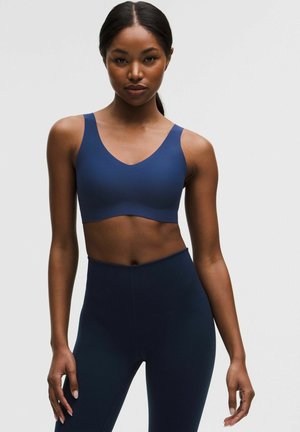 Femeie purtând un sutien sport și colanți skinny albastru închis asortați, stând pe un fundal simplu și deschis la culoare.