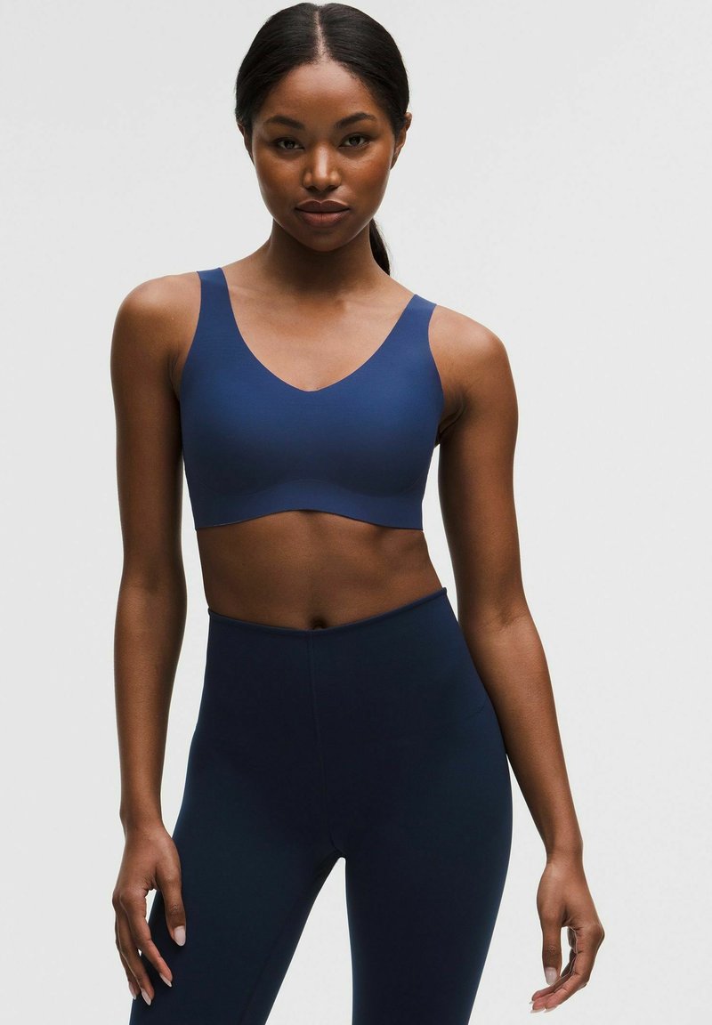 Frau, die einen passenden marineblauen Sport-BH und hoch taillierte Leggings trägt, steht vor einem einfarbigen hellen Hintergrund.