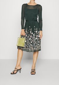 Dunkelgrünes Kleid mit langen durchsichtigen Ärmeln und floralen Stickereien am Saum. Hält eine kleine, strukturierte grüne Handtasche; trägt schwarze Riemchen-Heels.