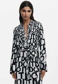 Schwarz-weißer Hemdkleid mit einem markanten, wiederholten "DON'T DO IT"-Grafik, langen Ärmeln und einem Gürtel zum Binden in der Taille. Glatte Stofftextur.