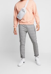Man wearing peach sweatshirt, gray plaid pants, white sneakers, and a light gray crossbody bag against a white studio background.