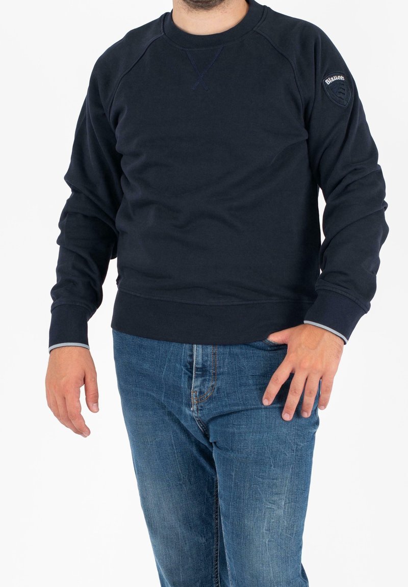 Homme portant un sweat-shirt bleu marine avec un écusson logo Blauer sur la manche gauche et un jean bleu, debout devant un fond blanc.