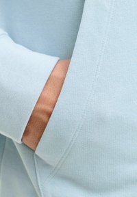 Main partiellement glissée dans la poche avant d'un sweat-shirt bleu clair avec des détails de couture visibles.