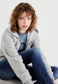 Siva zip-up dukserica s mekim teksturom, nosi se preko plave majice s grafikom. Model sjedi, nosi tamnoplave uske traperice.