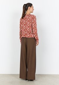 Rote Bluse mit Muster und langen Ärmeln, kombiniert mit weiten, braunen hoch taillierten Hosen. Glatte Stofftextur sichtbar.