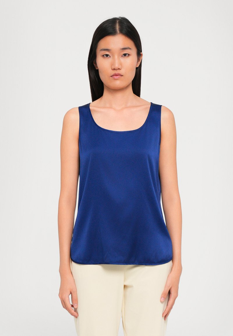 Woman with long black hair wearing a sleeveless navy blue satin top and light beige pants, standing against a plain light background.