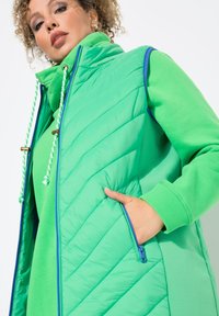 Chaleco aislante verde con diseño acolchado, cuello alto, bolsillo con cremallera, capucha ajustable con cordón decorativo y puños de canalé.