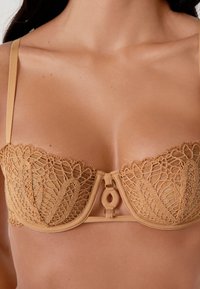 Primer plano de una mujer que lleva un sujetador balconette de encaje beige con tirantes finos y un cierre decorativo en la parte frontal.
