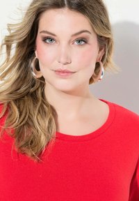 Maglione rosso a maglia con scollo rotondo. Capelli lunghi e mossi. Grandi orecchini a cerchio in argento. Sfondo neutro. Pelle chiara e trucco naturale.