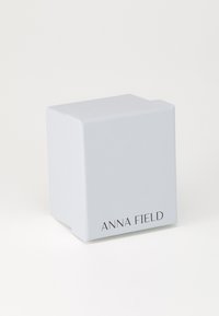 Hvid emballageæske med en glat tekstur, hvor mærkenavnet "ANNA FIELD" er trykt i mørke bogstaver på den ene side.