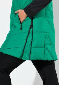Gilet puffer verde con texture a trapunta, tasche laterali con zip e colletto alto. Indossato sopra abiti neri.