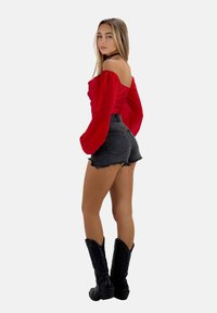 Top rosso scollato con spalle scoperte e maniche a sbuffo, abbinato a pantaloni shorts in denim nero a vita alta e stivali da cowboy neri con ricami decorativi.