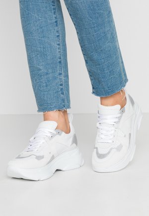 Person iført lyseblå cropped jeans og hvide chunky sneakers med grå accenter, stående på et hvidt gulv.