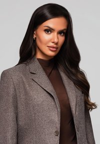 Hnědý herringbone blazer s jedním knoflíkem a texturovanou tkaninou, nošený přes tmavě hnědý top. Obsahuje klopový límec a kapsu.
