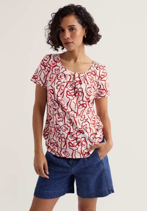 Kobieta o kręconych ciemnych włosach, ubrana w białą bluzkę z krótkim rękawem z czerwonym kwiatowym wzorem oraz niebieskie dżinsowe szorty, stojąca na jasnym tle.