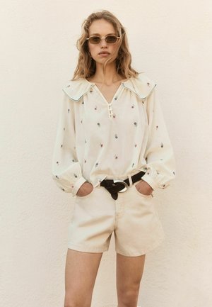 Tânără cu păr ondulat purtând ochelari de soare mici, o bluză crem cu imprimeuri florale, pantaloni scurți bej și o curea neagră, stând în fața unui perete deschis la culoare.