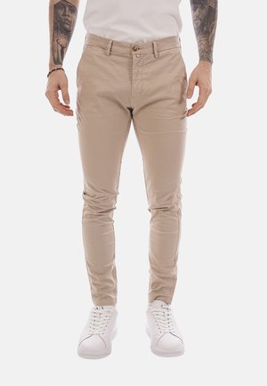 Uomo che indossa pantaloni chino beige slim-fit, sneaker bianche e una camicia bianca, con tatuaggi su tutto il braccio e un braccialetto nero al polso sinistro.