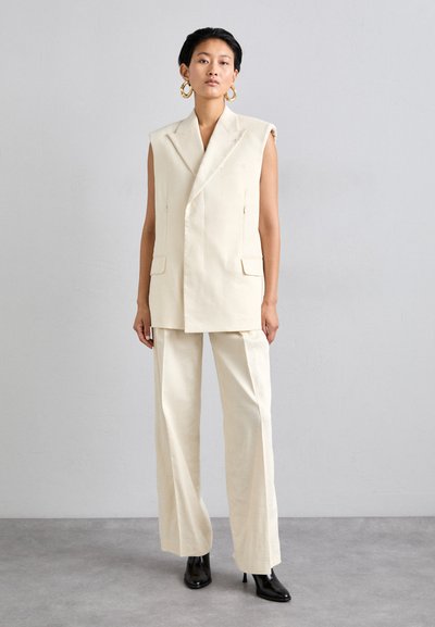 Veste sans manches en lin beige avec fermeture croisée à double bouton et deux poches, associée à un pantalon large et des bottes à talons noires.