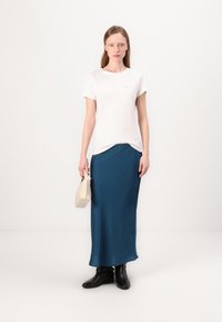 T-shirt bianco in cotone con un logo sottile, abbinato a una gonna lunga teal, stivali neri alla caviglia e una piccola borsa crema. Design semplice.