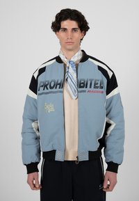 Homme portant une veste de course bleue et noire avec le texte « PROHIBITED RACING », un pull beige, un foulard bleu et blanc à motifs autour du cou, et un pantalon sombre.