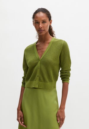 Femme portant un cardigan boutonné texturé vert olive à col en V et une jupe lisse assortie, debout sur un fond clair uni.