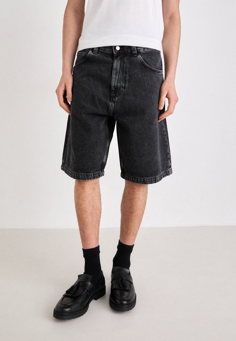 Schwarze Denim-Shorts mit lockerer Passform, ausgestattet mit einem klassischen Fünf-Taschen-Design und umgeschlagenem Saum. Kombiniert mit schwarzen Slippern und Crew-Socken.