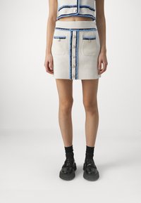 Witte mini-jurk met blauwe rand, voorzien van een knoopsluiting aan de voorkant en twee voorzakken. Gecombineerd met een bijpassend cropped top en zwarte schoenen.