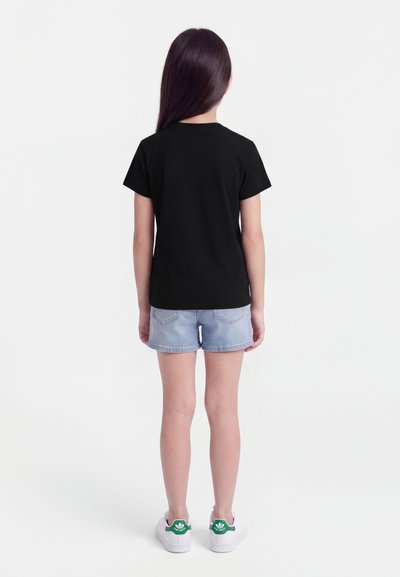 Otrok z dolgimi temnimi lasmi, ki nosi preprosto črno majico, svetle denim hlače in bele superge z zelenimi logotipi, stoji obrnjeno stran.