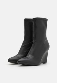 Bottines à talons compensés en cuir noir avec un bout pointu, texture lisse et un détail de couture minimal. Conçues pour une apparence élégante et ajustée.