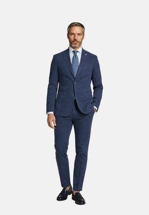 Uomo che indossa un abito blu navy con pantaloni coordinati, cravatta azzurra, camicia bianca e mocassini in pelle nera, in piedi con una mano in tasca.
