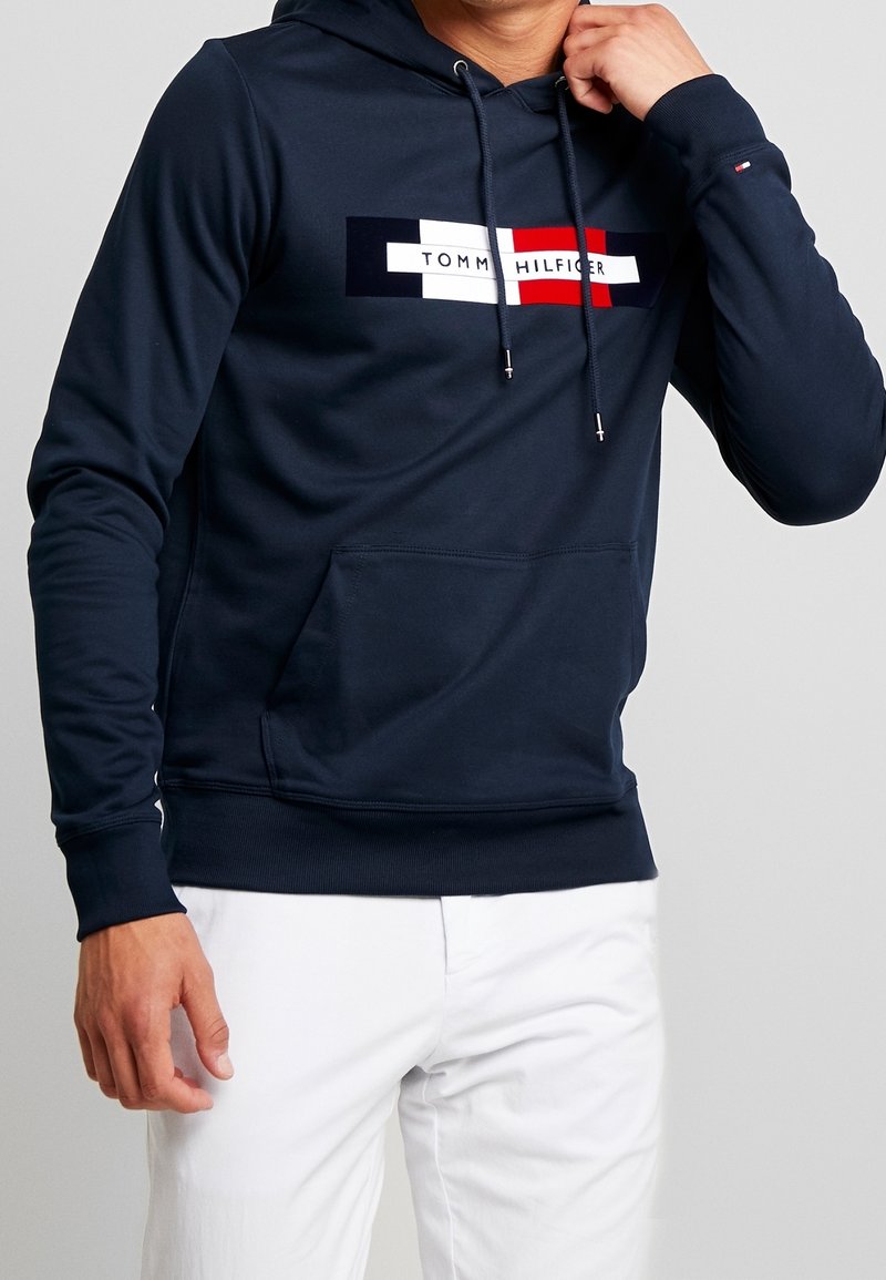 Granatowy hoodie wykonany z mieszanki bawełny. Posiada przednią kieszeń typu kangur, kaptur ze sznurkiem i logo w kolorze czerwonym, białym i niebieskim.