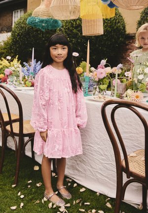 Jeune fille en robe fleurie rose se tient près d'une table extérieure dressée avec des fleurs, des bougies et des abat-jours tissés suspendus dans un cadre de jardin.