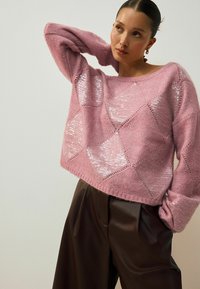 Różowy sweter dzianinowy z wzorem diamentowym, ozdobiony cekinami, zestawiony z brązowymi spodniami o wysokim stanie z plisami. Miękka tekstura, luźny krój.